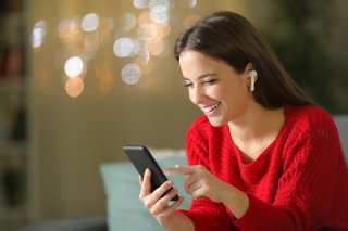 Happy girl in red listening to music with phone and wireless earbuds in the night sitting on a couch in the living room at home